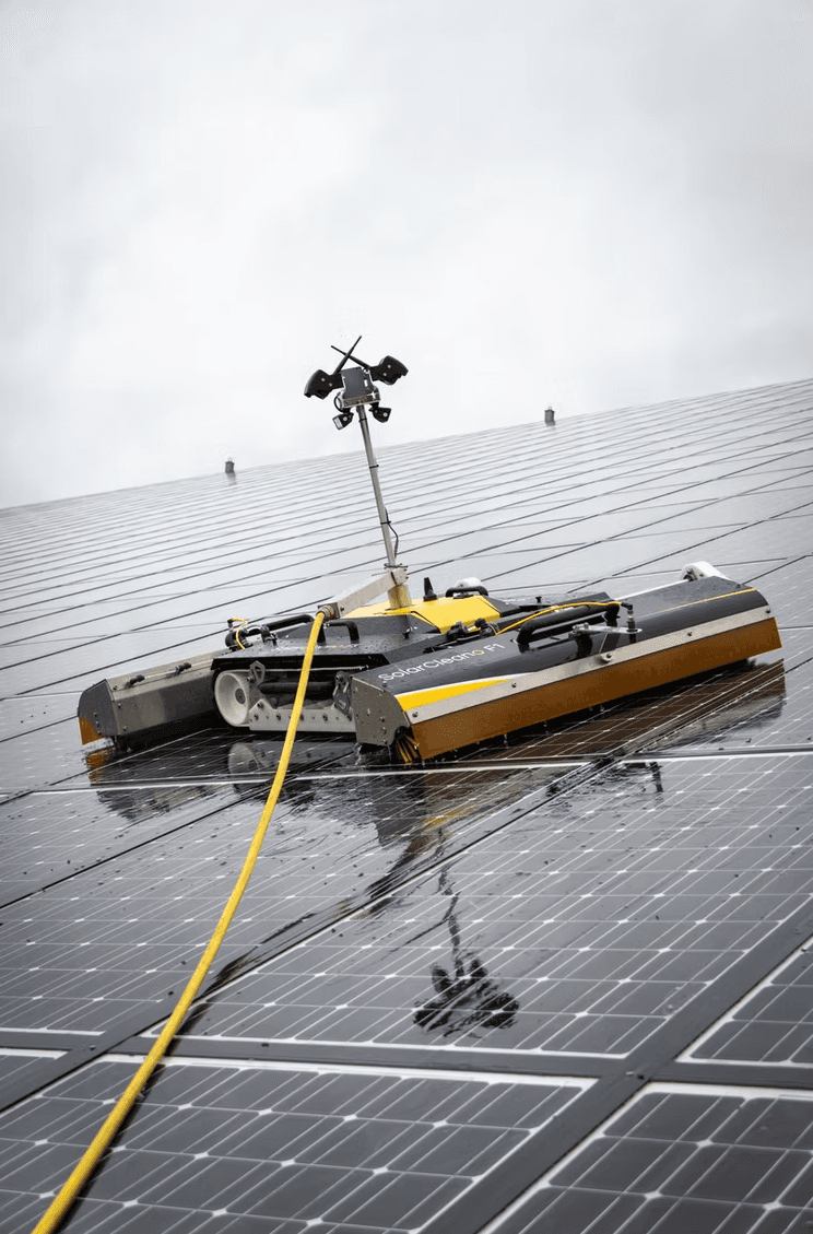 Robot SolarCleano F1 limpiando una gran superficie de paneles solares mojados con manguera amarilla.
